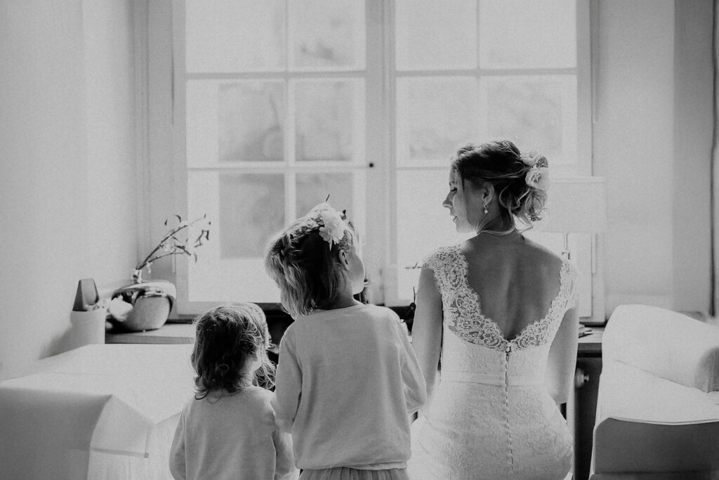 Braut im Spitzenkleid mit zwei Kindern blickt zum Fenster mit weichem Licht. Titel: Braut-kinder-zum-Fenster.
